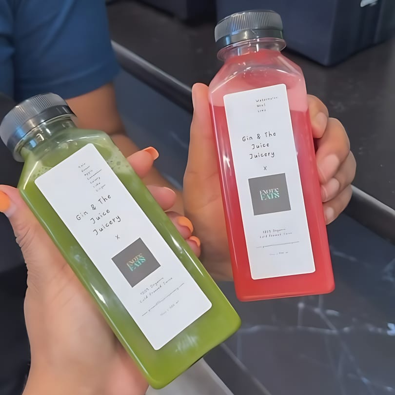 Two hands holding bottles of "Gin & The Juice Juicery" drinks, one green and one red.