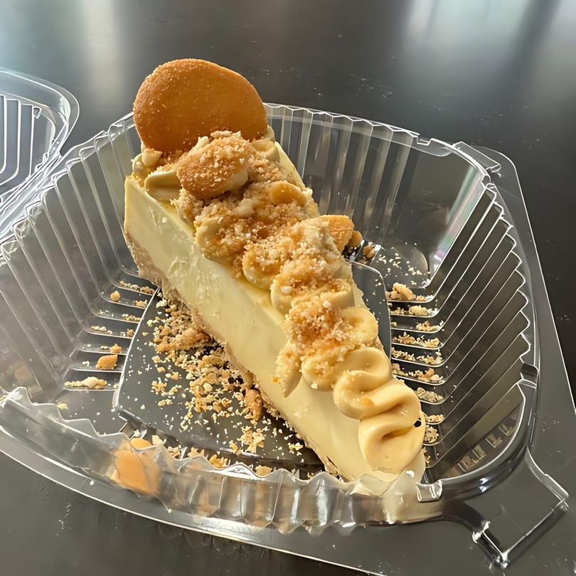 A slice of banana pudding cheesecake with cookie crumbles and a vanilla wafer in a plastic to-go container.