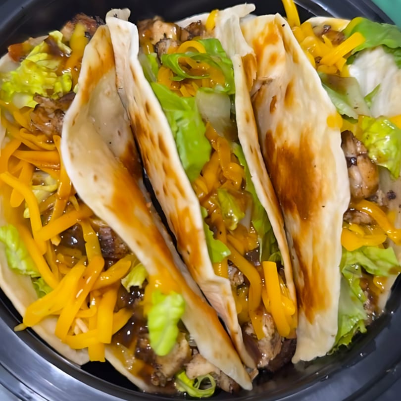 Three soul-food tacos filled with chicken, shredded cheese, lettuce, and sauce in a black container.