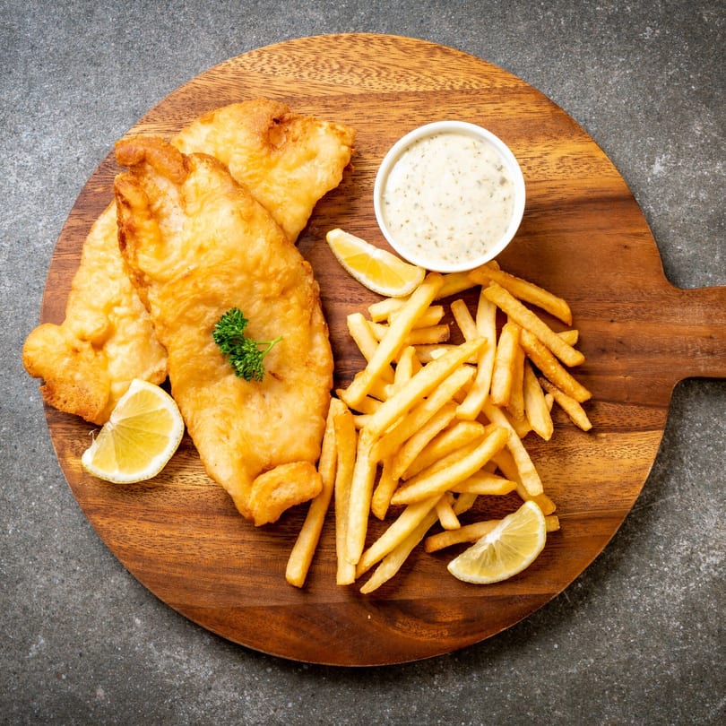 Golden fried fish and chips with tartar sauce and lemon wedges on a wooden board.