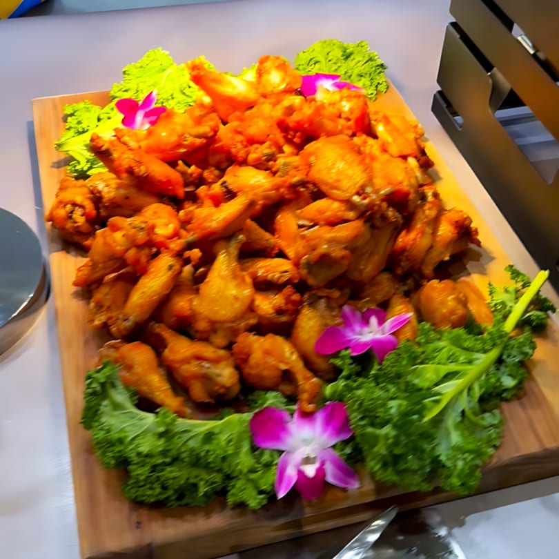 A large wooden platter filled with golden-brown fried chicken wings, garnished with lettuce and purple orchids.