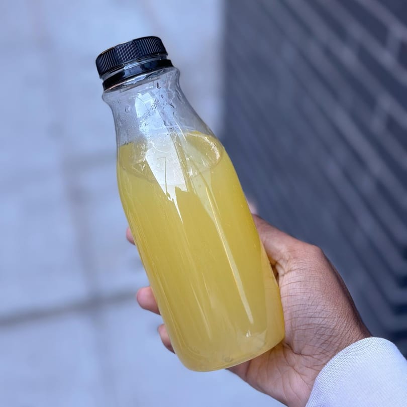 A hand holds a clear plastic bottle filled with a yellow beverage, possibly lemonade, from Exotic Eats NJ.