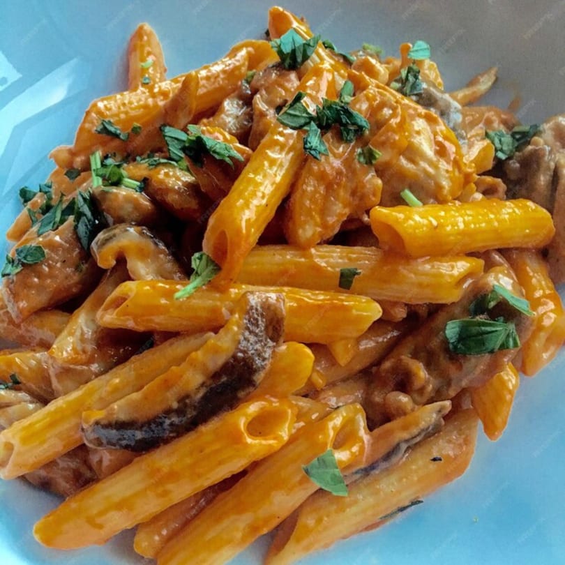 Penne pasta with creamy orange sauce, mushrooms, and fresh green herbs in a white bowl.