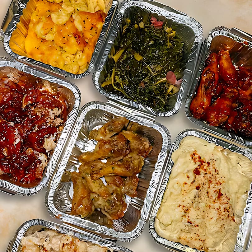 Assortment of soul food dishes in foil containers, including mac and cheese, collard greens, and chicken.